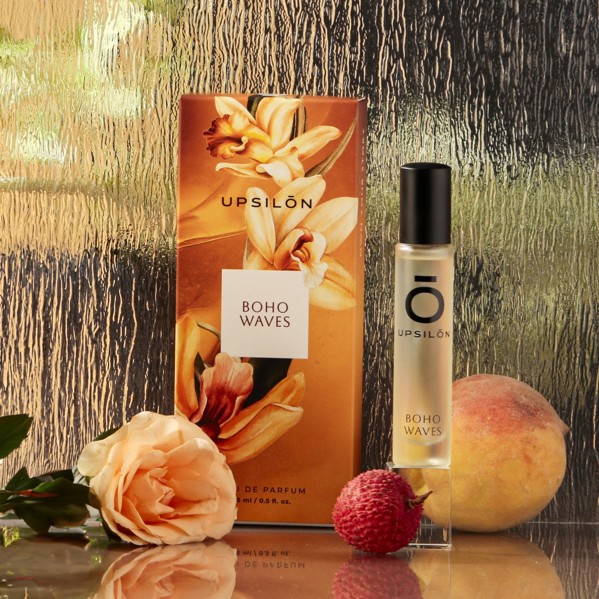 Upsilon Boho Waves Eau de Parfum bottle with a gold cap and pink label, surrounded by fresh flowers and colorful fruit.