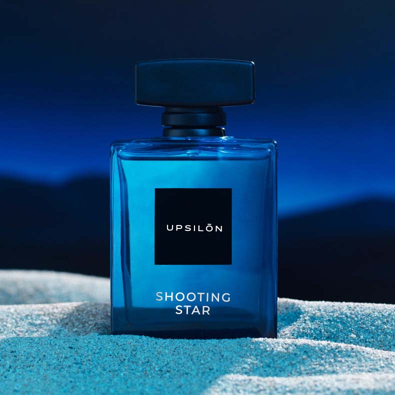 Upsilon Shooting Star Eau de Parfum sits atop a mound of shimmering gold sand,