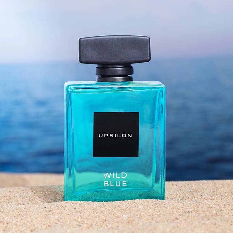 UPSILON Wild Blue Eau de Parfum bottle on a sandy beach.