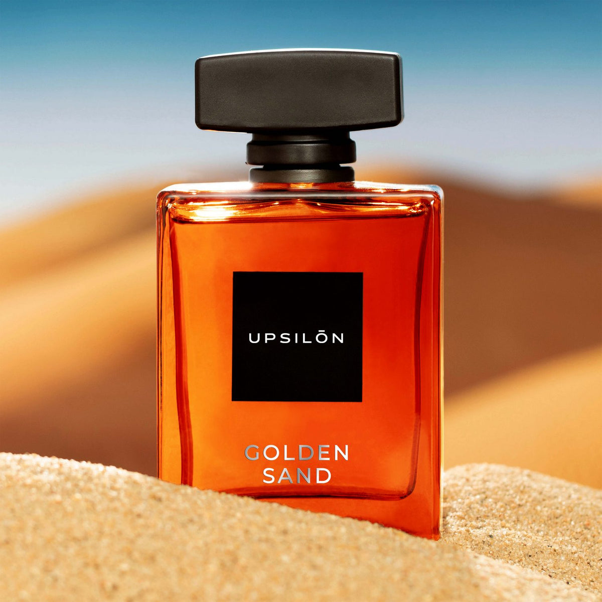 A bottle of UPSILON Golden Sand Eau de Parfum sitting on top of a pile of sand on a beach
