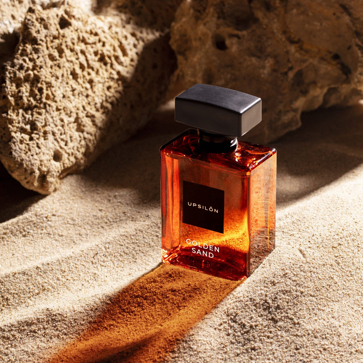 A bottle of UPSILON Golden Sand Eau de Parfum sits atop a sun-drenched pile of sand